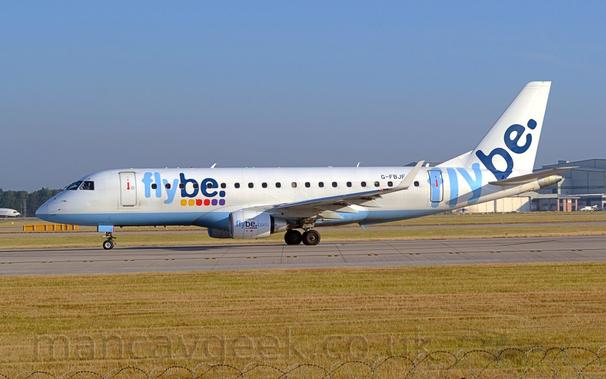 Side view of a white, twin engined jet airliner facing to the left on a grey runway.
The plane has a pale blue belly, and pale and dark blue "FlyBe" titles above a series of yellow, red, and purple dots on the forward fuselage.
These titles are repeated on the sides of the engine cowlings  with additional "[dot] com" titles, as well as on the tail, minus the dots.
Green grass fills the bottom of the frame, with the razor-wire on the top of the perimeter fence visible right at the bottom.
More grass in the background leads up to a large grey hangar in the distance on the right, and trees on the left, under blue-grey sky.