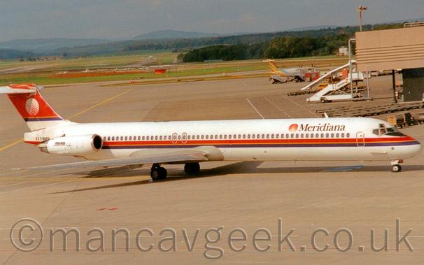 Side view of a white, twin engined jet airliner with the engines mounted on the sides of the rear fuselage, taxiing from left to right.
The plane has a red and purple stripe running along the body just below the passenger cabin windows, with yellow piping down the middle.
There are black "Meridiana" titles on the upper forward fuselage, next to a red and white circular logo.
The tail is mostly red, with a purple stripe and yellow piping at the bottom, with the circular logo repeated, only this time with the colours inverted.
The registration "I-SMES" is on the tail-cone, behind the engines, next to an Italian flag.
The engines have small, black "MD-82" titles.
Grey concrete apron fills the foreground, and a good chunk of the background, with a sandy-brown terminal building just edging it's way in to shot on the right.
A white, high-winged, twin propellor-engined airliner with yellow stripes on the tail is parked behind a brown prefabricated hut and a couple of sets of mobile airstairs in the background.
Some light aircraft are parked on grass in the distance on the left, with more grass and trees beyond that leading off to the skyline, under hazy grey sky.