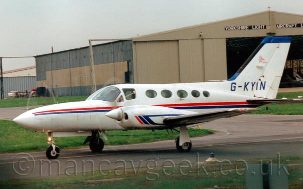 Side view of a white, twin engined BizProp taxiing from right to left.
The plane has a red and blue stripe running along the body, and short, red and blue diagonal bars on the engine cowlings, the latter of which sweeps back to go over and under the wings.
The registration "G-KYIN" is on the tail in blue.
Green grass lines both sides of the taxiway the plane is on, with a large brown and grey hangar with the doors wide open in the background.
Black boards over the hangar doors have the words "Yorkshire Light Aircraft" in large white letters.
Grey sky fills the rest of the frame.