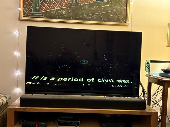 A television displays scrolling text against a starry background, stating, "It is a period of civil war." The scene is set in a cozy living room with ambient lighting, a patterned sofa, and a small table with a gaming console.