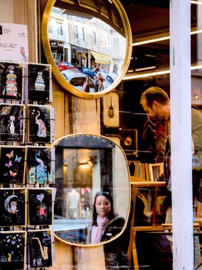 The image shows the interior of a shop with two mirrors reflecting the street scene outside. The upper mirror displays cars and buildings, while the lower mirror shows a woman standing outside. To the left, there are shelves with various greeting cards featuring colorful designs. Inside the shop, a man appears to be browsing items. The overall scene captures a blend of reflections and products.