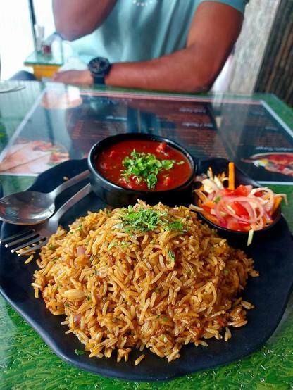 A plate of fried rice, a bowl of red sauce, and a small salad on a black plate. A fork and spoon are beside the rice.