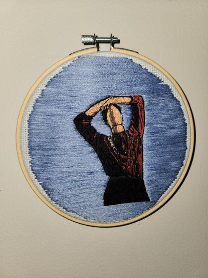 An embroidery project of BTS's Jin, standing against a blue background with his arms resting on the top of his head. He's wearing a burgundy sweater, black pants, and silver jewelry, and he's partly in shadow.