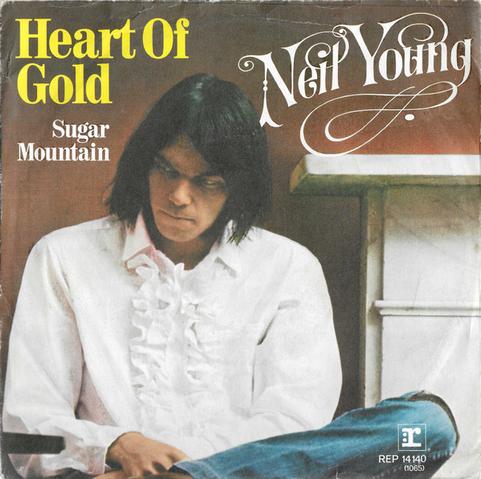 Cover art for Neil Young's single 'Heart of Gold' featuring Neil Young seated in a white ruffled shirt and jeans, looking down pensively beside a fireplace. Text includes 'Heart of Gold', 'Sugar Mountain', and 'Neil Young' in stylized fonts