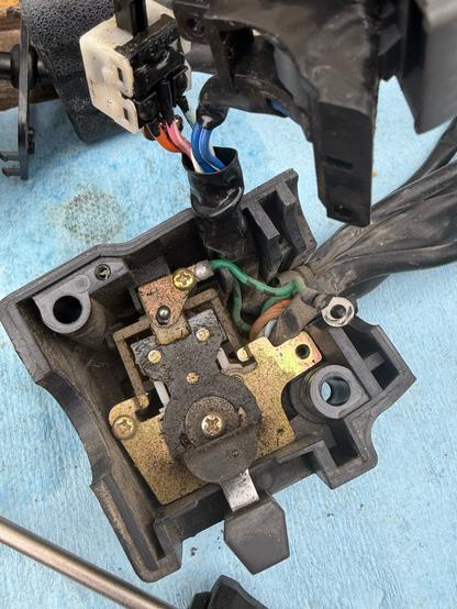 the inside of a motorcycle turn signal hand controls. there are many small screws, brass plates, and wires connected in random places, with a lever sticking out of its black plastic housing. it is very greasy and dirty.