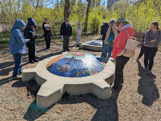 A group of people standing around a sculpture in a public park. The sculpture is a concrete turtle on the ground. It's back is a large mosaic of various images, including a black Thunderbird prominently visible in the photo. A second turtle sculpture can be seen further back.

This piece is called mamohkamatowin (Helping Each Other) by Jerry Whitehead.