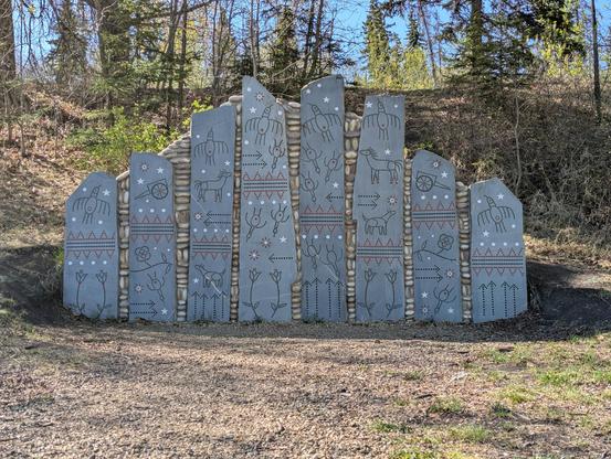 A sculpture in a public park of 8 large stone slabs standing to against a wall of river stones. There slabs have images in the style of petroglyphs of birds, flowers, horses, carts, and other imagery.

Preparing to Cross the Sacred River by Marianne Nicolson