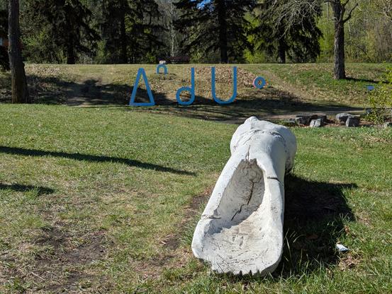 Two sculptures in a public park.

In the foreground is a large white sculpture made to look like an oversized bone hide scraper. This is mikikwan by Duane Linklater.

In behind are large Cree syllabics built onto a small hill- ᐃᐢᑯᑌᐤ -  iskotew by Amy Malbeuf