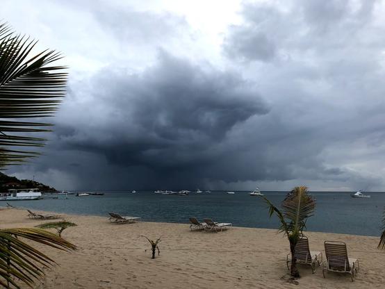 Stretch of sand with a few reclining beach chairs reaches toward a dark blue sea above which the clouds form a curious face over looking the water. The clouds are dark, and where eyes would be there are two lighter spots, and another suggesting a mouth. Above what could be the face, the clouds lighten making the figure appear to be wearing an acorn shaped hat. Dark streaks of clouds stretch out from either side as if arms are opened wide. Surely rain is falling where the clouds meet the sky.