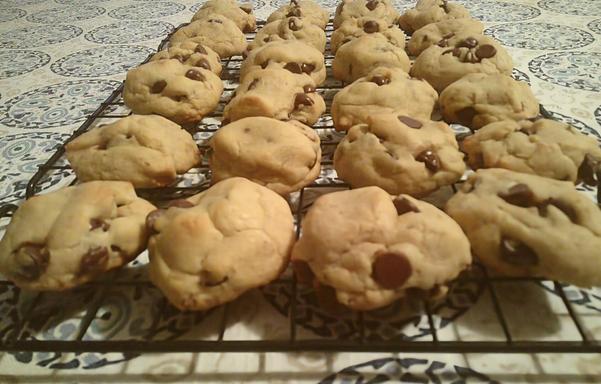 Numerous freshly baked chocolate chip cookies are arranged on a black wire cooling rack. The cookies are a light golden brown and generously studded with melted chocolate chips.