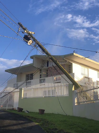 A wooden electric pole topple over at about a 45 degree angle, leaning against a concrete and metal fence. Several power lines are pulled and down and a cylindrical transformer box hangs from the top of the pole.