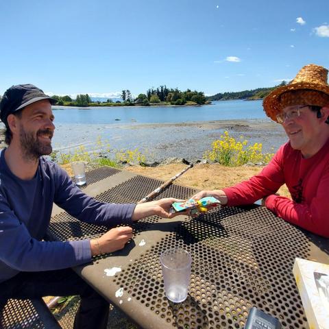 Two men are sitting at a weathered metal picnic table outdoors, exchanging a small, colorfully wrapped gift. The man on the left, with a beard and a black cap, smiles as he accepts the gift. The man on the right, wearing a cedar bark woven hat and glasses, also smiles. A body of water with islands is in the background, with yellow wildflowers growing along the grassy edge. The scene is bathed in sunlight on a clear, bright day.