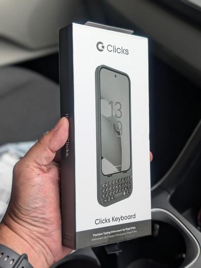 A person holds a box for a Clicks Keyboard, a device designed for the Pixel 9 Pro phone. The box features an image of the keyboard attached to a smartphone, and text indicating its purpose as a precision typing instrument. The packaging is seen from a side angle and held by the person's hand.