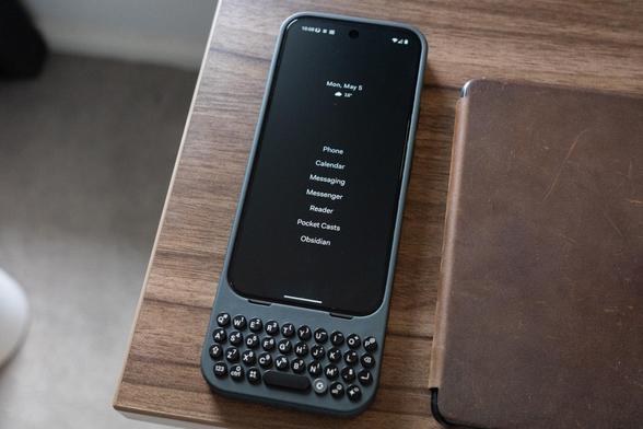 A smartphone with a physical keyboard attachment is lying on a wooden table. The screen displays a simple black and white interface with the date \"Mon, May 5\" at the top, followed by a list of apps: Phone, Calendar, Messaging, Messenger, Reader, Pocket Casts, and Obsidian. Beside the phone is a brown leather-bound notebook or wallet.
