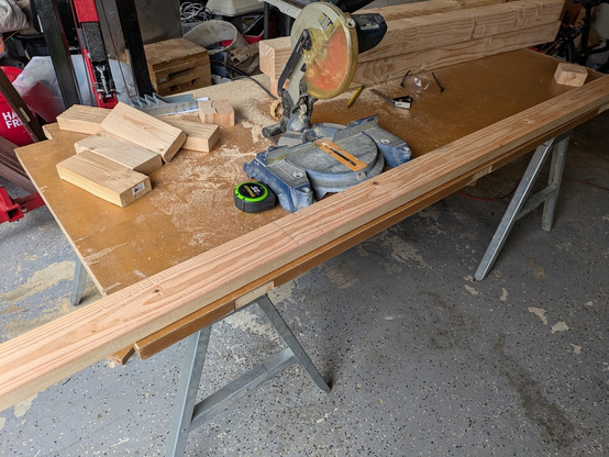 A workbench in progress. A door sits on a pair of sawhorses and supports a chop saw, measuring tools, and several pieces of cut lumber.