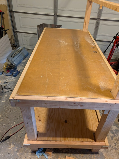 The workbench partially assembled. The door now sits in a frame of 2x4 lumber supported by 4x4 posts. A plywood shelf is assembled below the main bench.