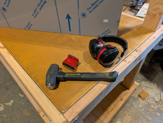 A piece of stainless steel sits on the partially-assembled workbench, next to a pair of ear protectors, a sledgehammer and a dolly block.