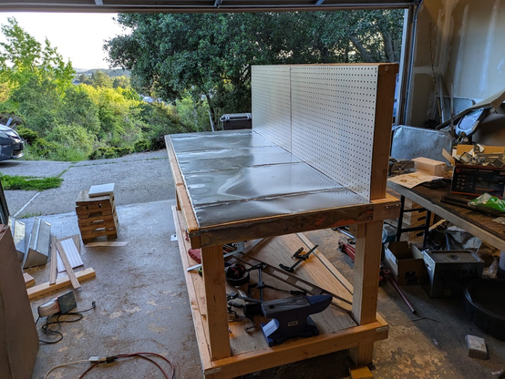 The workbench, now completed, sits in a pile of sawdust. The top surface is now covered by somewhat wrinkly stainless steel, there is a pegboard at the back of the bench, and numerous tools including an anvil sit on the lower shelf.