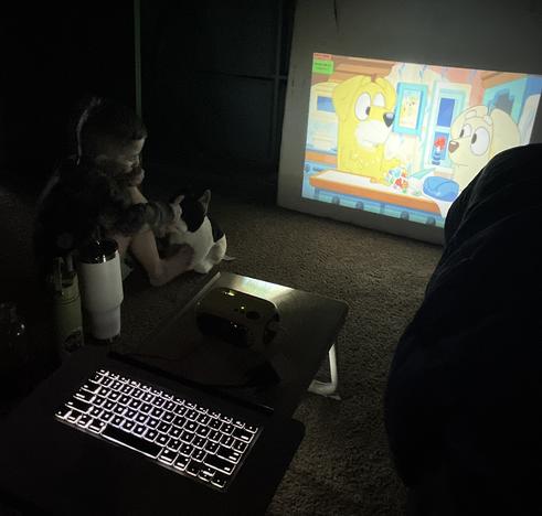 Dimly lit room with a kiddo watching Bluey projected on the wall  from a cheap projector and laptop that is missing a screen.