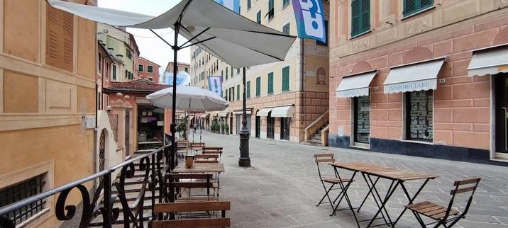 A charming European street with tables, chairs, and umbrellas outside a cafe. Buildings with colorful facades line the street.