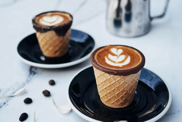 Two coffee cups made of waffle cones, with chocolate rims, on black saucers. A metal pitcher is in the background.
