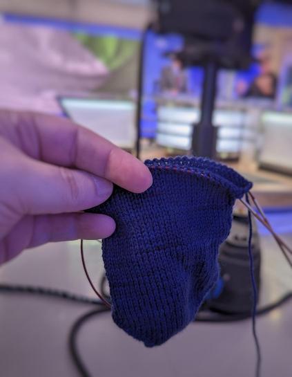 Close up of a sock in progress of being knit. In the background is a blurry television camera and set. There are two guys sitting on the set but their heads are obscured by the camera.