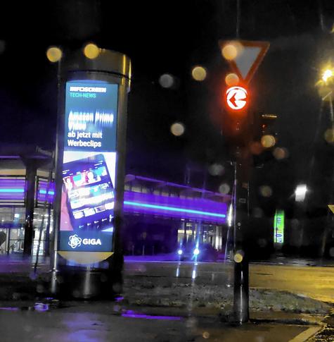 Nachts, direkt neben einer Ampel: eine über 4m hohe Display-Säule mit "Infoscreen Tech-News: Amazon Prime Video ab jetzt mit Werbeclips" - einem Bild dazu und "GIGA" als "Nachrichten"-Verbreiter. Es ist etwas Regen auf der Scheibe.