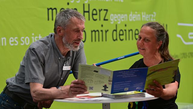 Im Vordergrund an einem Stehtisch der Standbesucher links und die Pastorin rechts. Sie hält einen blauen Stab in ihrer rechten Hand. Beide sehen gemeinsam in ein Faltblatt, das sie gemeinsam halten. Im Hintergrund der verkürzte Bibelvers.
