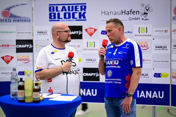 Daniel R. Schmidt, links, interviewt den Fanbeauftragten der HSG Hanau vor einer Sponsorenwand.