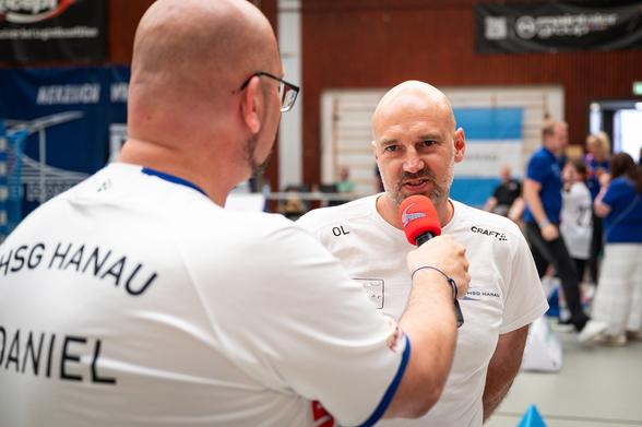 Im unscharfen Vordergrund ist der Rücken von Hallensprecher Daniel R. Schmidt zu sehen, der gerade den im Brennpunkt des Fotos befindlichen Oli Lücke - Co-Trainer der HSG Hanau - am Mikrofon hat.