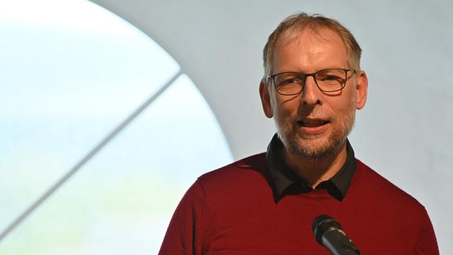 Porträt von Frank Aichele. Ein Mannes mittleren Alters mit kurzen, grauen Haaren und einer Brille. Er trägt einen dunkelroten Pullover und spricht in ein Mikrofon. Der Hintergrund ist unscharf und zeigt einen hellen Raum mit einem Teil eines großen Fensters.
