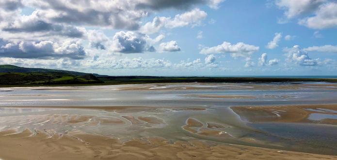 This picture showcases a serene coastal landscape with vast sandy tidal flats, gently interspersed with shallow pools of water reflecting the sky. The horizon reveals a distant stretch of green land with softly rolling hills. Above, the sky is partly cloudy, filled with fluffy white clouds contrasting against a bright blue backdrop. The sunlight casts a warm glow on the sand, accentuating the natural patterns formed by the receding tide. The overall scene exudes tranquility and natural beauty, likely captured during low tide.