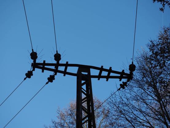 The image shows a silhouette of a power line pole against a clear blue sky. The pole has several insulators and power lines extending from it. There are also trees visible on both sides of the pole.