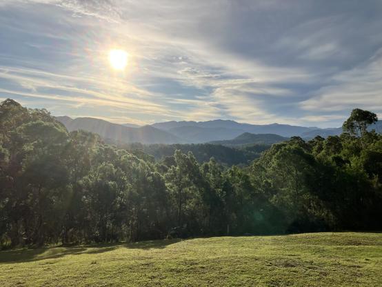 The sun sits low in the sky over the Brogo River Valley. Mountains lie in the distance with nearby forest visible over a cleared grassy area.