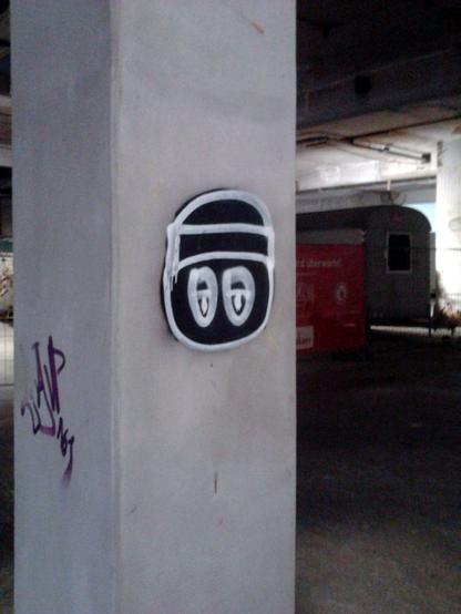 Graffito: Eine schnell gesprühte, gesichtsähnliche Form auf einem Betonpfeiler im Sockelgeschoss der Ruine des Ihmezentrums in Hannover-Linden. Im Hintergrund unscharf erkennbar: Ein Bauwagen und ein Werbetransparent an einer Baustellenabsperrung.