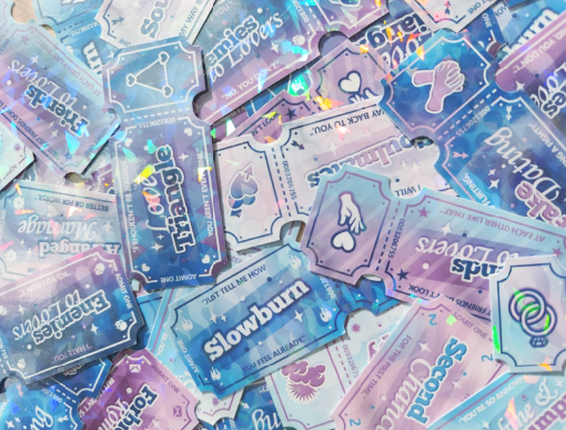 photo of my many stickers shaped like tickets and they have romance tropes written on them such as slowburn, love triangle, enemies to lovers etc.