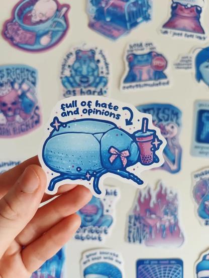 hand holding up a sticker of a blue bug with ribbon titled "full of hate and opinions"