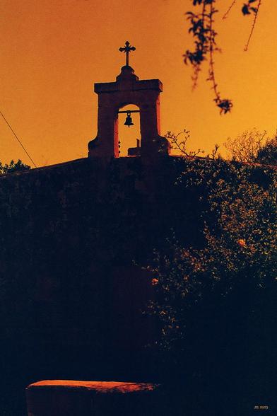 Farbfoto mit auf Rotskala reduziertem Farbspektrum, das eine Frontalansicht einer alten Kirche in Zejtun auf Malta zeigt, von deren Fassada sich nur das Glockenportal mit einer Glocke und Kreuz obenauf abhebt. Rechts grünes und oranges Gesprenkel eines Busches, oben rechts herabhängende Zweige eines Baums. Links unten ist die sonnenbeschienene Oberfläche einer Steinbank erkennbar. Leica R5 mit 50 mm Summilux auf Lomography Redscale XR-Film.