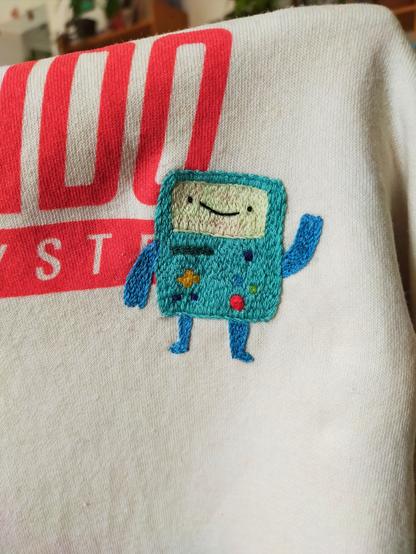 Bmo (une console de jeu type gameboy anthropomorphe) brodée sur un sweat blanc Nintendo