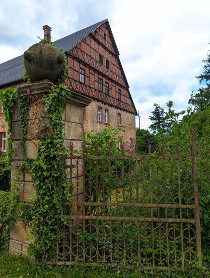A photograph shows a building with a red brick and dark roof. The building's side is visible, featuring multiple windows. An ornate stone gatepost, covered in ivy, stands in the foreground to the left. A wrought-iron fence runs behind the gatepost, partially obscured by greenery. The sky is overcast with light clouds.