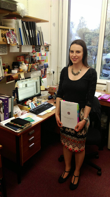 A photo of the poster, Emma Hodcroft, on 5 May 2015. She is wearing a dark blue top and orange-and-blue patterned skirt, and looking nervous but smiling. She holds a very thick copy of her thesis (printed one-sided and double-spaced as regulations required) with many coloured page-marking tabs sticking out of the sides & top. She stands before a wooden desk covered in notes, notebooks, post-its, small knick-nacks and toys (and a computer), in Ashworth Building at University of Edinburgh, where she did her PhD.