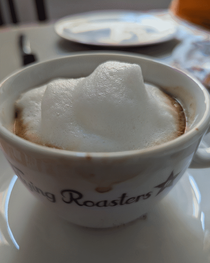 Nahaufnahme einer Kaffeetasse (eigentlich Cappucchino), die mit Kaffee und einer hoch aufgeschäumten Menge Milchschaum gefüllt ist. Tasse steht auf einer Untertasse, im leichten Gegenlicht, wohl auf einem Küchentisch. Im Hintergrund ist ein leerer Teller und etwas in orange zu sehen. Auf der Tasse steht in lilan Schreibschrift Flying Roasters geschrieben.