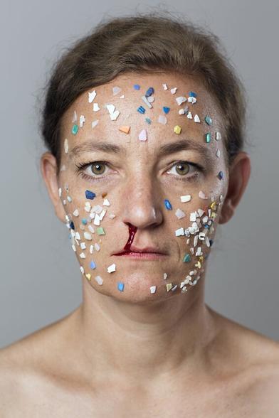 Photographic portrait of the conceptual artist Swaantje Güntzel. The picture shows the artist's face covered with different colored microplastic particles. Blood flows from the artist's right nostril. Swaantje Güntzel looks directly into the camera and wears her brown hair pinned back, her neck and parts of her shoulders are uncovered. The microplastic particles were collected during an intervention at Playa de Montaña Bermeja in Lanzarote where she cleared 1 squaremeter of microplastics. 1370 pieces were counted.