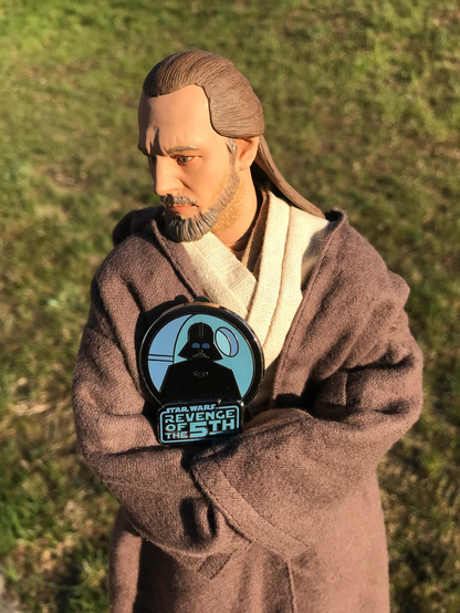 A Qui-Gon figure, standing outside against a backdrop of green grass, holds a sign with an image of Darth Vader that says, “Star Wars: Revenge of the 5th.”