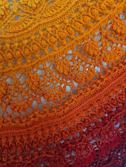 A close-up of a crocheted shawl showcasing its intricate stitch patterns. The fabric features a gradient color transition from yellow at the top through orange and deep red at the bottom. The crochet work includes decorative motifs, such as leaf-like shapes and textured bobble stitches, highlighting the craftsmanship behind the design.