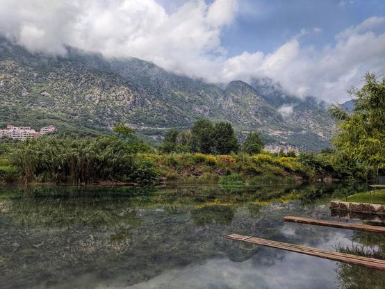 The image shows a tranquil landscape with a body of water, which is part of the Bay of Kotor, reflecting the surrounding scenery. The water is mostly still, creating a mirror-like effect of the sky and the lush vegetation on the banks. The sky is partly cloudy, with the clouds casting soft shadows on the water's surface. In the background, a mountain range rises, partially obscured by mist or low-hanging clouds, adding depth to the scene. The vegetation along the water's edge includes various trees and shrubs. Two wooden planks extend into the water from the near bank.