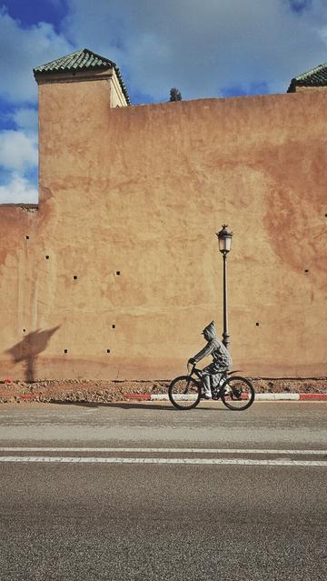 The portrait-orientated image shows a person on a bicycle riding past a wall. The person is wearing a traditional Moroccan hooded garment jelaba. There is a street lamp visible. The wall is made of light brown material and has a green tiled roof section. The ground is paved with asphalt and includes white lines marking lanes. There is a striped red line painted on the ground near the base of the wall, marking the beginning of the pavement. The sky is partly cloudy. Shadows are cast on the wall and the ground.