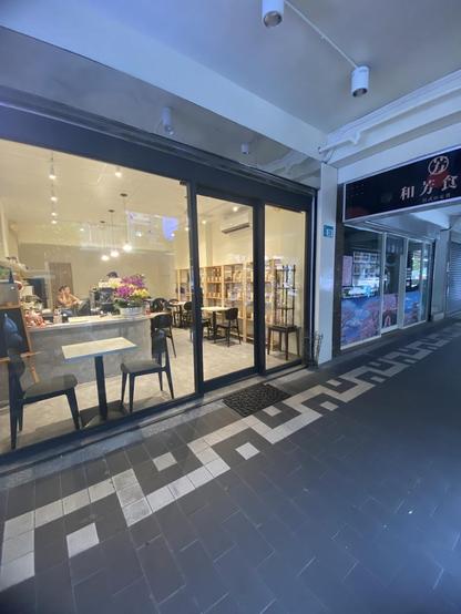 An exterior shot of two businesses. One business appears to be a cafe with tables and chairs visible through the large glass windows. Next to it is a restaurant with a sign in Japanese and images of Japanese scenery on the windows. The storefronts are adjacent to each other along a walkway with a patterned gray brick floor.