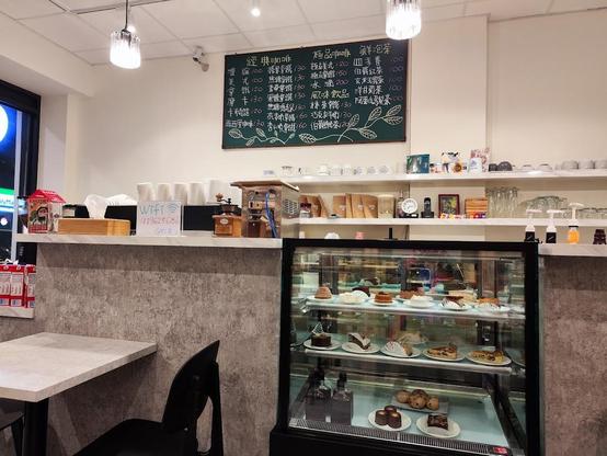 A cozy café scene with a chalkboard menu, a pastry display case filled with cakes and desserts, and a seating area with a table and chair. A countertop with a coffee grinder and a sign for "WiFi" adds to the inviting ambiance.