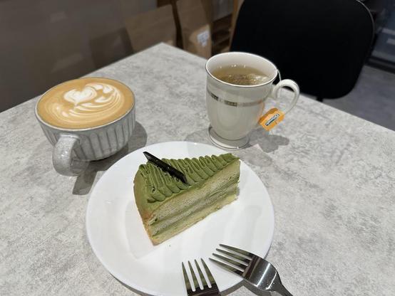 A table with a cup of coffee, a mug of tea, and a slice of green tea cake with a fork on a white plate.